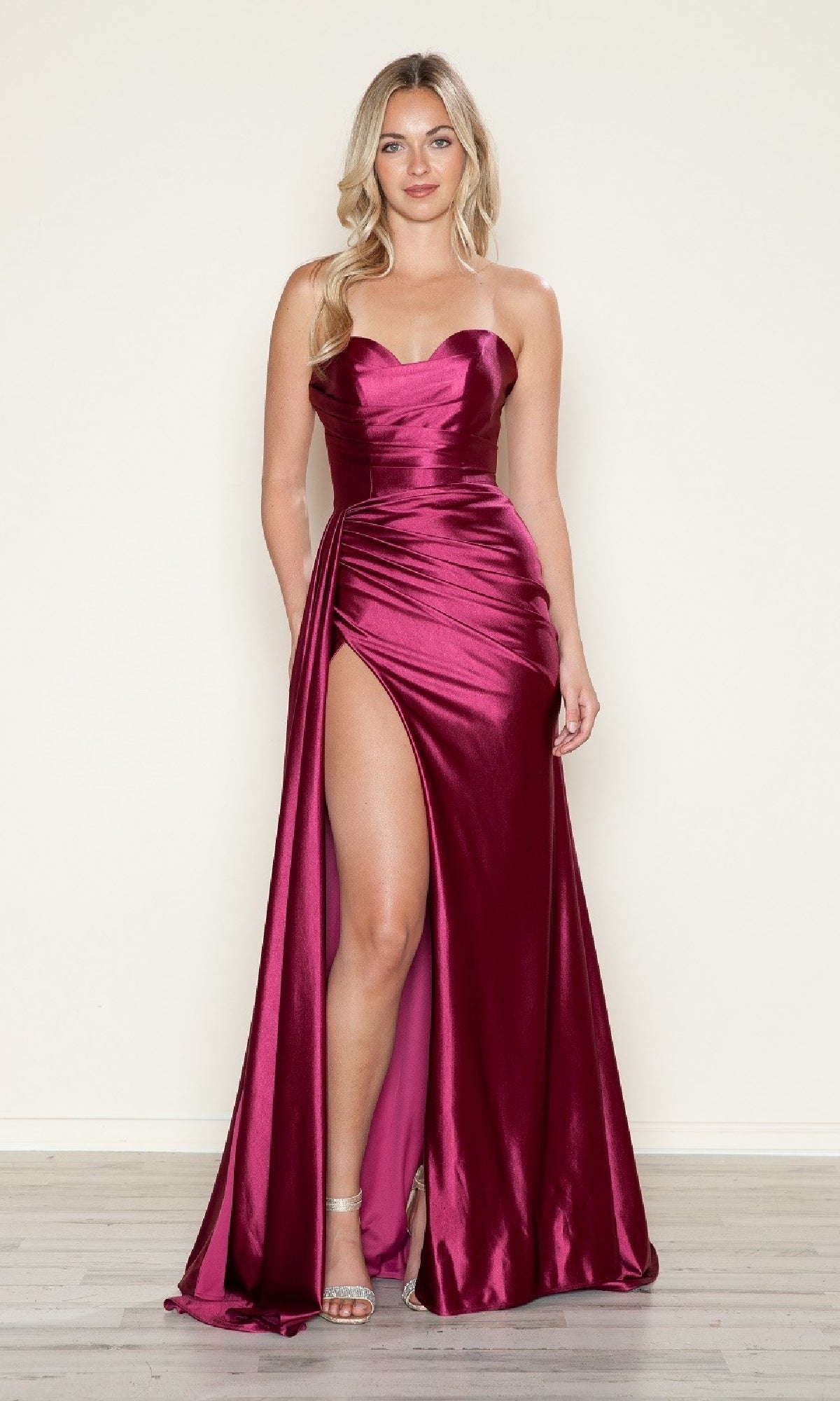 A woman in the Strapless Side-Sash Long Prom Gown (Poly USA 9734)—a magenta, strapless, floor-length satin dress with a thigh-high slit—stands on a light wood floor against a beige background. This elegant gown is under $200.